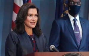 MI Governor Gretchen Whitmer speaking at a podium with a masked person standing to her right