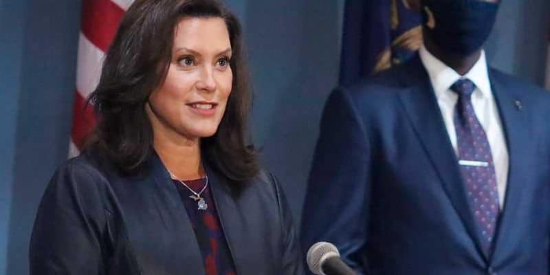 MI Governor Gretchen Whitmer speaking at a podium with a masked person standing to her right