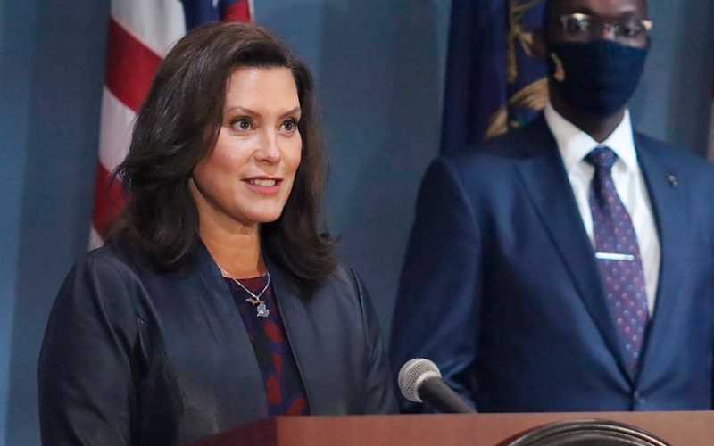 MI Governor Gretchen Whitmer speaking at a podium with a masked person standing to her right
